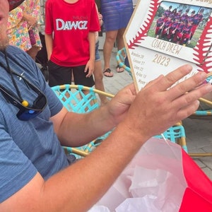 May include: A baseball-themed plaque with a photo of a team in red and black uniforms. The plaque reads "Polk County Dawgz COACH DREW 2023" with player names listed below. The plaque is held by a person.