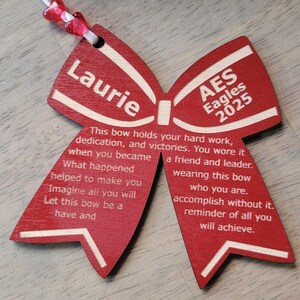 May include: A red wooden bow with white lettering. The bow has the name "Laurie" on one side and "AES Eagles 2025" on the other side. The text reads: "This bow holds your hard work, dedication, and victories. You wore it when you became a friend and leader. What happened helped to make you wearing this bow who you are. Imagine all you will accomplish without it. Let this bow be a reminder of all you will achieve."
