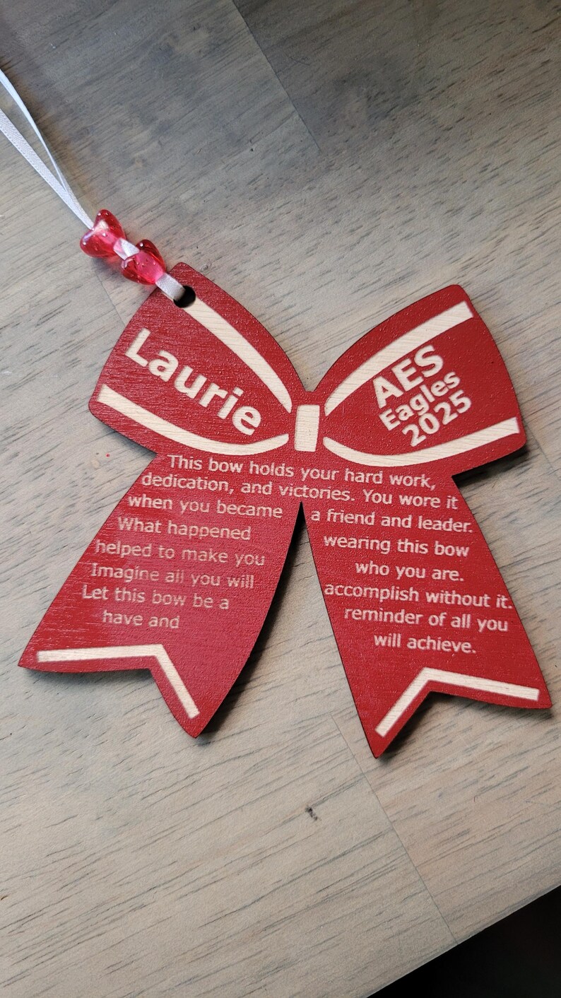 May include: A red wooden bow with white lettering. The bow has the name "Laurie" on one side and "AES Eagles 2025" on the other side. The text reads: "This bow holds your hard work, dedication, and victories. You wore it when you became a friend and leader. What happened helped to make you wearing this bow who you are. Imagine all you will accomplish without it. Let this bow be a reminder of all you will achieve."