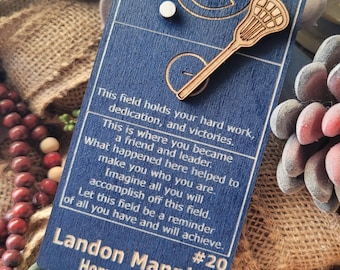 May include: A blue lacrosse-themed graduation tag with the text "Class of 2026" and a wooden lacrosse stick. The tag includes a personalized message and the name "Landon Manning" and "Homer Trojans".