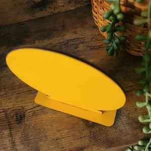 May include: A bright yellow oval-shaped wooden sign blank with a smooth, solid-colored surface. The oval piece is securely slotted into a matching rectangular yellow wooden base, allowing the sign to stand upright on a flat surface.