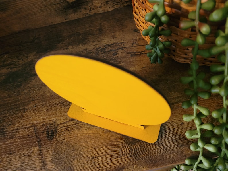 May include: A bright yellow oval-shaped wooden sign blank with a smooth, solid-colored surface. The oval piece is securely slotted into a matching rectangular yellow wooden base, allowing the sign to stand upright on a flat surface.