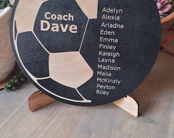 Puede incluir: Una placa circular de madera negra y natural con un diseño de balón de fútbol cortado con láser. El texto dice 2025 ASA Elite, Coach Dave, y una lista de nombres que incluye a Adelyn, Alexia, Ariadne, Eden, Emma, Finley, Kaleigh, Layna, Madison, Malia, McKinzly, Peyton y Riley.