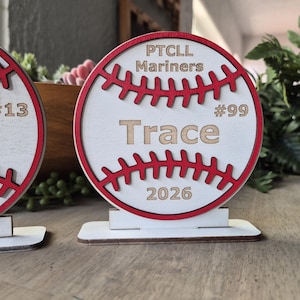 May include: A circular wooden baseball-shaped sign with a white face, red stitched detailing, and a red border. The text reads PTCLL Mariners, Trace, #99, and 2026. The sign is mounted on a rectangular white wooden base for upright display.