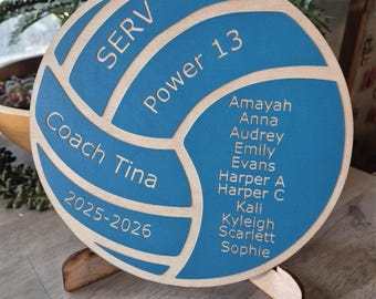 Könnte beinhalten: Eine kreisförmige hölzerne Volleyball-Plakette mit blau bemalten Paneelen und naturfarbenen Holzakzenten. Der Text lautet SERV, Power 13, Coach Tina, 2025-2026 sowie eine Namensliste: Amayah, Anna, Audrey, Emily, Evans, Harper A, Harper C, Kali, Kyleigh, Scarlett und Sophie.