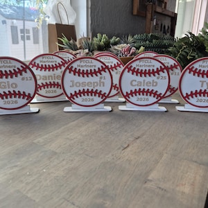 May include: A collection of circular wooden baseball-themed plaques, each featuring red stitched detailing, the text PTCL Mariners, a player name, a jersey number, and the year 2026. Each plaque is mounted on a small rectangular white base.