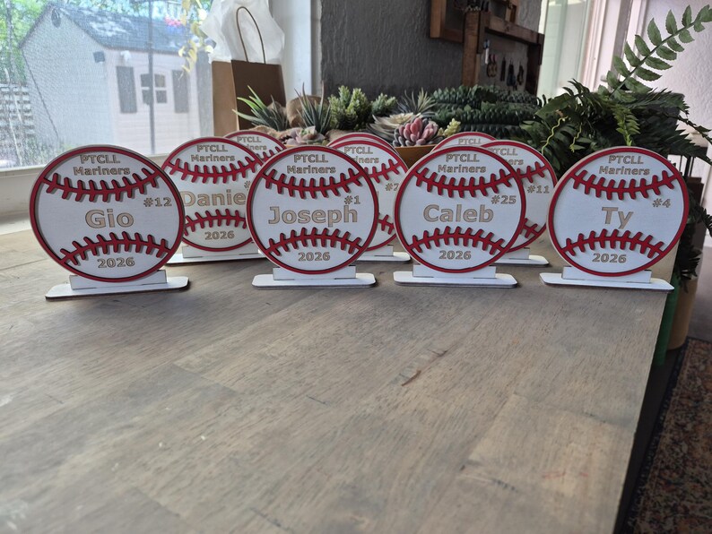 May include: A collection of circular wooden baseball-themed plaques, each featuring red stitched detailing, the text PTCL Mariners, a player name, a jersey number, and the year 2026. Each plaque is mounted on a small rectangular white base.