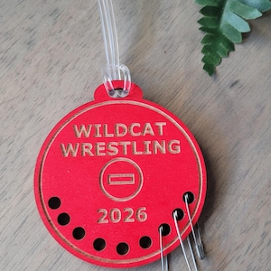 May include: A red wooden ornament with the words "WILDCAT WRESTLING" and the year "2026" in gold lettering. The ornament has a clear plastic loop for hanging and several safety pins attached. The background includes greenery.