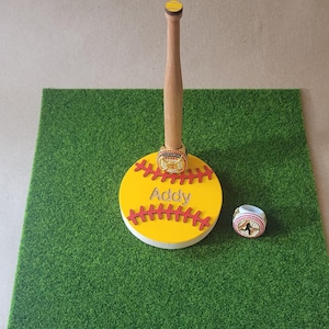 May include: A wooden baseball bat display stand featuring a yellow circular base with red stitching and the name Addy in silver lettering. A gold championship ring sits on the bat, and a second ring with a softball graphic sits nearby.