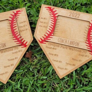 May include: Two wooden baseball-themed plaques with a baseball stitch design and a photo frame. The plaques are engraved with the year 2022, team names, and player names. The plaques are on a bed of green grass.