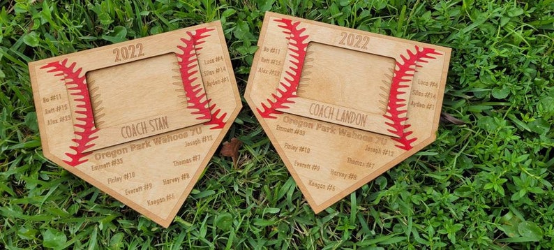 May include: Two wooden baseball-themed plaques with a baseball stitch design and a photo frame. The plaques are engraved with the year 2022, team names, and player names. The plaques are on a bed of green grass.