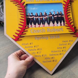May include: A wooden softball-themed photo frame in yellow and red, featuring a team photo and player names. The frame reads "Red Wolves 10U" and "Coach Robert, Spring 2026". Player names and numbers are listed below the photo.