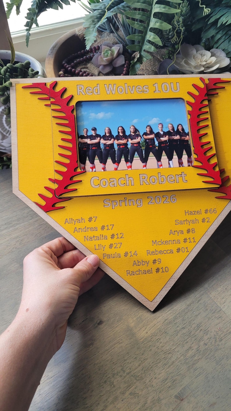 May include: A wooden softball-themed photo frame in yellow and red, featuring a team photo and player names. The frame reads "Red Wolves 10U" and "Coach Robert, Spring 2026". Player names and numbers are listed below the photo.