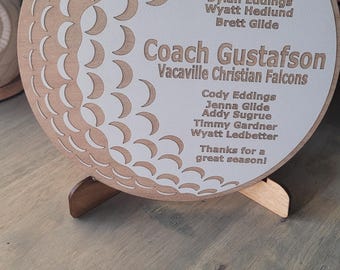 May include: A wooden plaque with a golf ball design and the text "2025 Coach Gustafson Vacaville Christian Falcons" and a list of names. The plaque is on a wooden stand.
