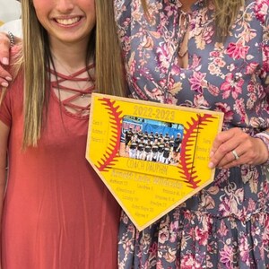 May include: A wooden plaque in the shape of a baseball home plate. The plaque has a yellow background with red stitching and the text "2022-2023". A team photo is in the center, with names listed on the sides. The text "Coach Dauphin" is also visible.