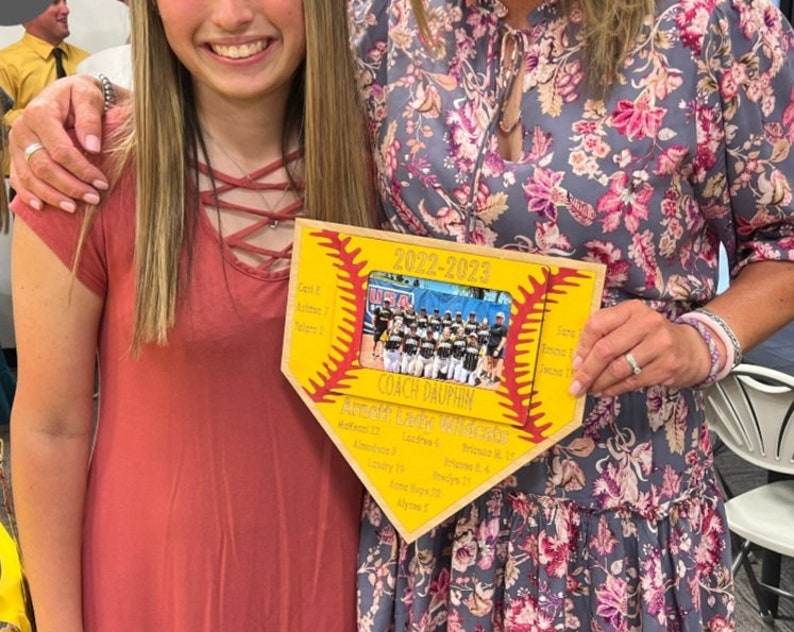 May include: A wooden plaque in the shape of a baseball home plate. The plaque has a yellow background with red stitching and the text "2022-2023". A team photo is in the center, with names listed on the sides. The text "Coach Dauphin" is also visible.