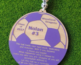 May include: A round wooden ornament shaped like a soccer ball with purple accents. It features the text Woodland Dragons, Class of 2027, Nolan #3, and an inspirational quote. The ornament hangs from a white cord with two small soccer ball beads.