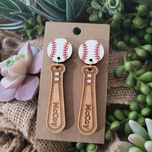 Puede incluir: Un par de pendientes colgantes con una pelota de b&eacute;isbol blanca con costuras rojas unida a un bate de b&eacute;isbol de madera marr&oacute;n claro. El bate tiene el nombre McCoy grabado verticalmente en la parte frontal. Presentados en una tarjeta.