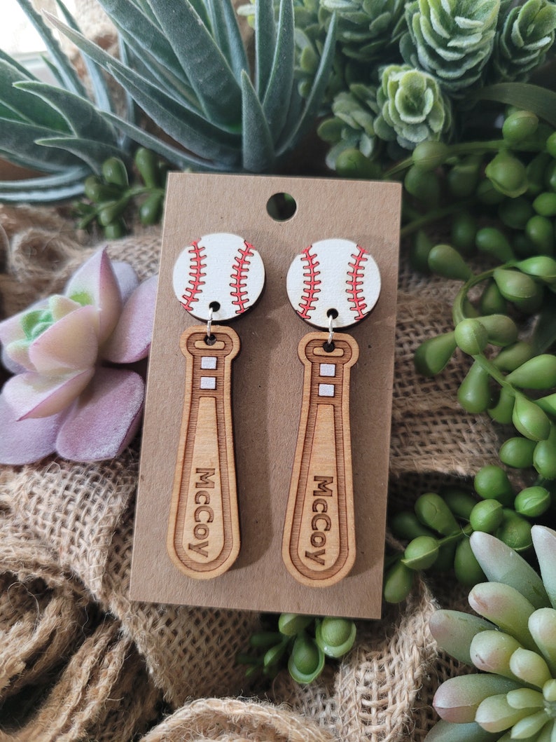 Puede incluir: Un par de pendientes colgantes con una pelota de b&eacute;isbol blanca con costuras rojas unida a un bate de b&eacute;isbol de madera marr&oacute;n claro. El bate tiene el nombre McCoy grabado verticalmente en la parte frontal. Presentados en una tarjeta.