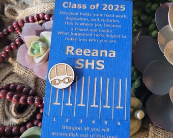 May include: A rectangular blue wooden ornament with a white ribbon, featuring the text Class of 2025, Reena SHS, and an inspirational message about a pool. It includes a small wooden swimming cap icon and lane markings numbered one through six.