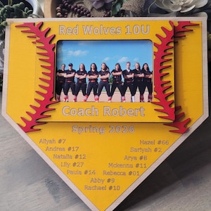 May include: A yellow home plate-shaped wooden picture frame with red baseball stitching accents. It features the text Red Wolves 10U, Coach Robert, Spring 2026, and a list of player names and numbers. A photograph of a softball team is centered.