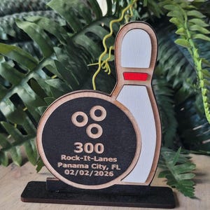 May include: A wooden bowling-themed trophy featuring a black circular bowling ball with three finger holes and a white bowling pin with a red stripe. The text reads 300, Rock-It-Lanes, Panama City, FL, and 02/02/2026 on a black base.