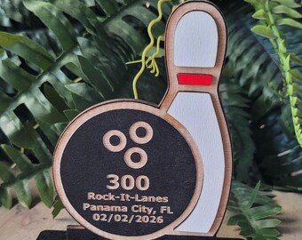 May include: A wooden bowling-themed trophy featuring a black circular bowling ball with three finger holes and a white bowling pin with a red stripe. The text reads 300, Rock-It-Lanes, Panama City, FL, and 02/02/2026 on a black base.