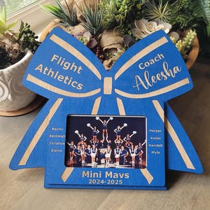 May include: A blue wooden picture frame shaped like a cheer bow. The frame features the text Flight Athletics, Coach Aleesha, Mini Mavs, 2024-2025, and a list of names. A photograph of a cheerleading team is displayed in the center of the frame.
