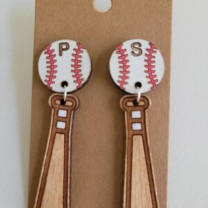 Puede incluir: Un par de pendientes colgantes con un dije de bate de b&eacute;isbol de madera unido a un dije circular de pelota de b&eacute;isbol. Las pelotas son blancas con costuras rojas y tienen las letras P y S en negro. Los bates son de madera color marr&oacute;n claro.