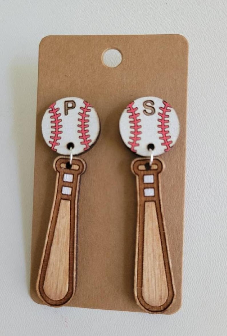 Puede incluir: Un par de pendientes colgantes con un dije de bate de b&eacute;isbol de madera unido a un dije circular de pelota de b&eacute;isbol. Las pelotas son blancas con costuras rojas y tienen las letras P y S en negro. Los bates son de madera color marr&oacute;n claro.