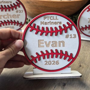 May include: A circular wooden baseball-themed sign with a white face, red stitched detailing, and a flat base. The text reads PTCLL Mariners, #13, Evan, and 2026. The design features layered wood elements and is displayed alongside similar items.