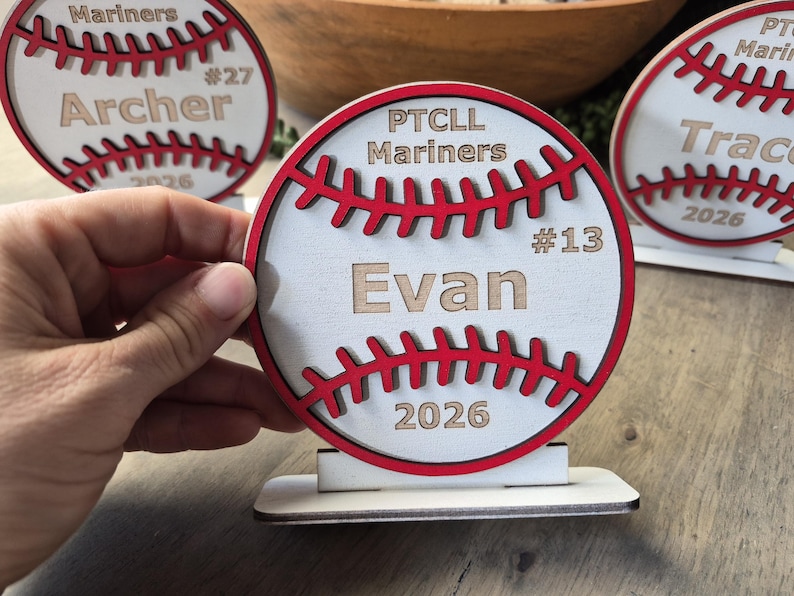May include: A circular wooden baseball-themed sign with a white face, red stitched detailing, and a flat base. The text reads PTCLL Mariners, #13, Evan, and 2026. The design features layered wood elements and is displayed alongside similar items.