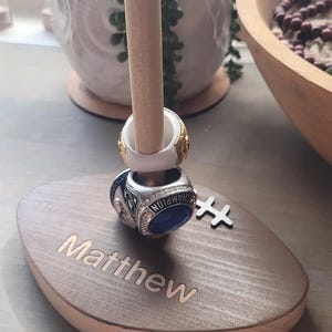 Op de afbeelding: Een houten ringhouder met een verticale deuvel die twee ringen ondersteunt. De ovale basis is gegraveerd met de naam "Matthew". De ringen zijn goud en zilver met blauwe en witte accenten. Rechts is een klein houten accent.