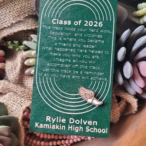 May include: A green wooden tag with white text that reads "Class of 2026" and a message about hard work and accomplishments. The tag also includes the name "Rylie Dolven" and "Kamiakin High School". A small wooden winged foot is attached.
