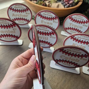 May include: A collection of circular wooden baseball-themed ornaments, each featuring red stitched details, the text PTCL Mariners, a player name, a jersey number, and the year 2026. Each ornament is mounted on a small white rectangular base.