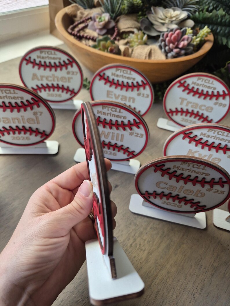 May include: A collection of circular wooden baseball-themed ornaments, each featuring red stitched details, the text PTCL Mariners, a player name, a jersey number, and the year 2026. Each ornament is mounted on a small white rectangular base.