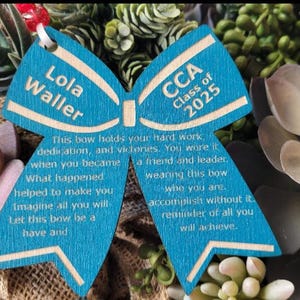 May include: A blue wooden bow with the text "Lola Waller" and "CCA Class of 2025" on it. The bow has a message about hard work, dedication, and victories.