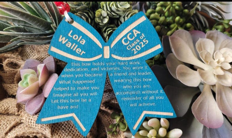 May include: A blue wooden bow with the text "Lola Waller" and "CCA Class of 2025" on it. The bow has a message about hard work, dedication, and victories.