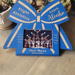May include: A blue wooden picture frame shaped like a bow with the words "Flight Athletics" and "Coach Aleesha" on it. The frame has a photo of a cheerleading squad with the words "Mini Mavs 2024-2025" below the photo. The names of the cheerleaders are listed on the left and right sides of the photo.