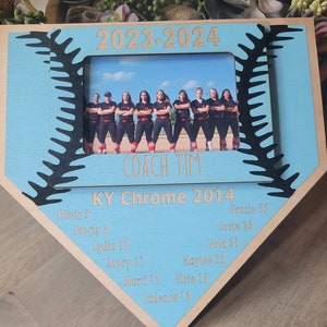 May include: A wooden baseball-shaped photo frame with a light blue center and a photo of a team. The frame includes the text "2023-2024", "COACH TIM", "KY Chrome 2014", and player names and numbers. The frame has a baseball stitch design.