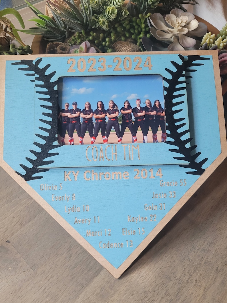 May include: A wooden baseball-shaped photo frame with a light blue center and a photo of a team. The frame includes the text "2023-2024", "COACH TIM", "KY Chrome 2014", and player names and numbers. The frame has a baseball stitch design.