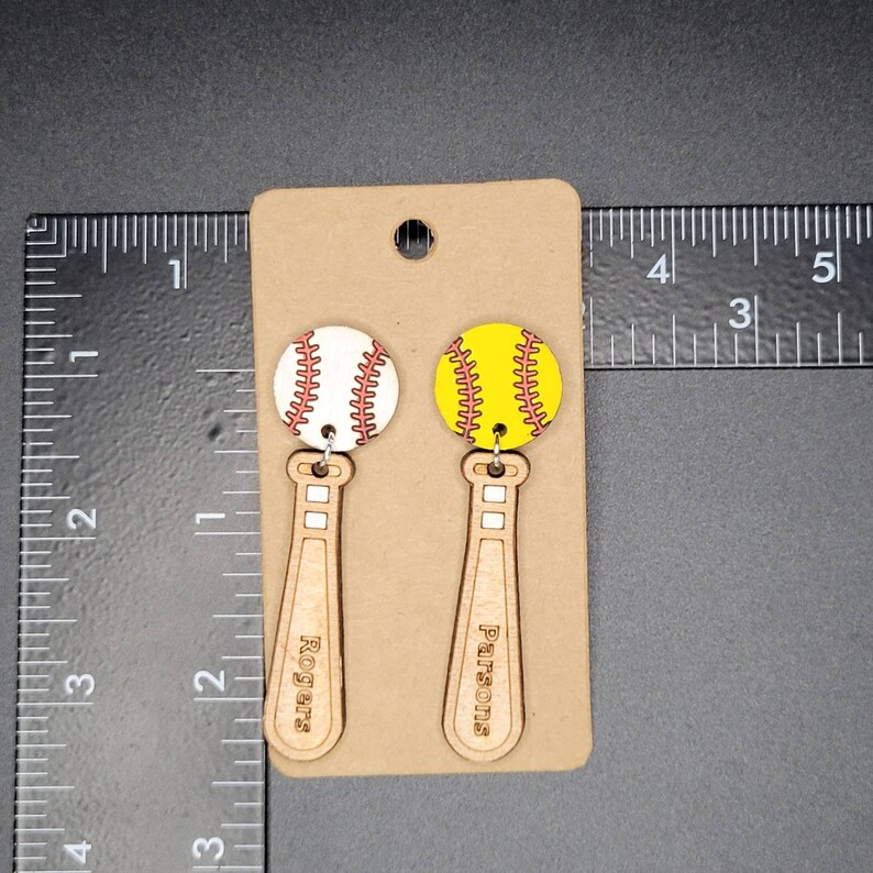 Puede incluir: Un par de pendientes colgantes con una pelota de b&eacute;isbol blanca y una de s&oacute;ftbol amarilla unidas a bates de b&eacute;isbol de madera. Los bates tienen grabados los nombres Rogers y Parsons. Una regla negra muestra una longitud de 5,08 cm.