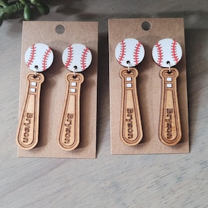 Puede incluir: Dos pares de pendientes colgantes con una pelota de b&eacute;isbol blanca con costuras rojas unida a un bate de b&eacute;isbol de madera marr&oacute;n claro. Cada bate tiene grabado el nombre Bryson en orientaci&oacute;n vertical sobre tarjetas de color marr&oacute;n.