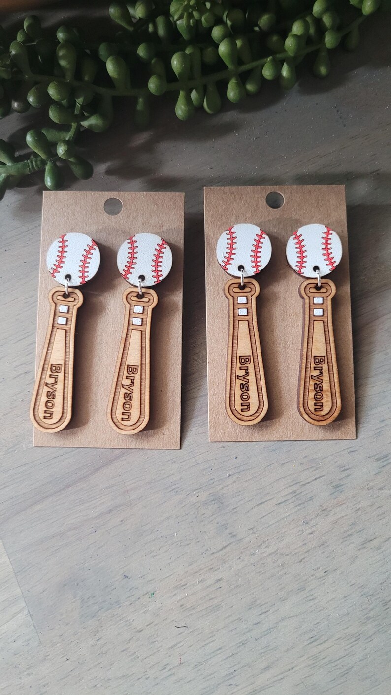Puede incluir: Dos pares de pendientes colgantes con una pelota de b&eacute;isbol blanca con costuras rojas unida a un bate de b&eacute;isbol de madera marr&oacute;n claro. Cada bate tiene grabado el nombre Bryson en orientaci&oacute;n vertical sobre tarjetas de color marr&oacute;n.