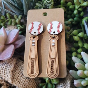 Puede incluir: Un par de pendientes colgantes con una pelota de b&eacute;isbol blanca con costuras rojas unida a un bate de b&eacute;isbol de madera marr&oacute;n claro. El nombre Dreessen est&aacute; grabado en negro en el bate. Los pendientes se presentan en una tarjeta beige.