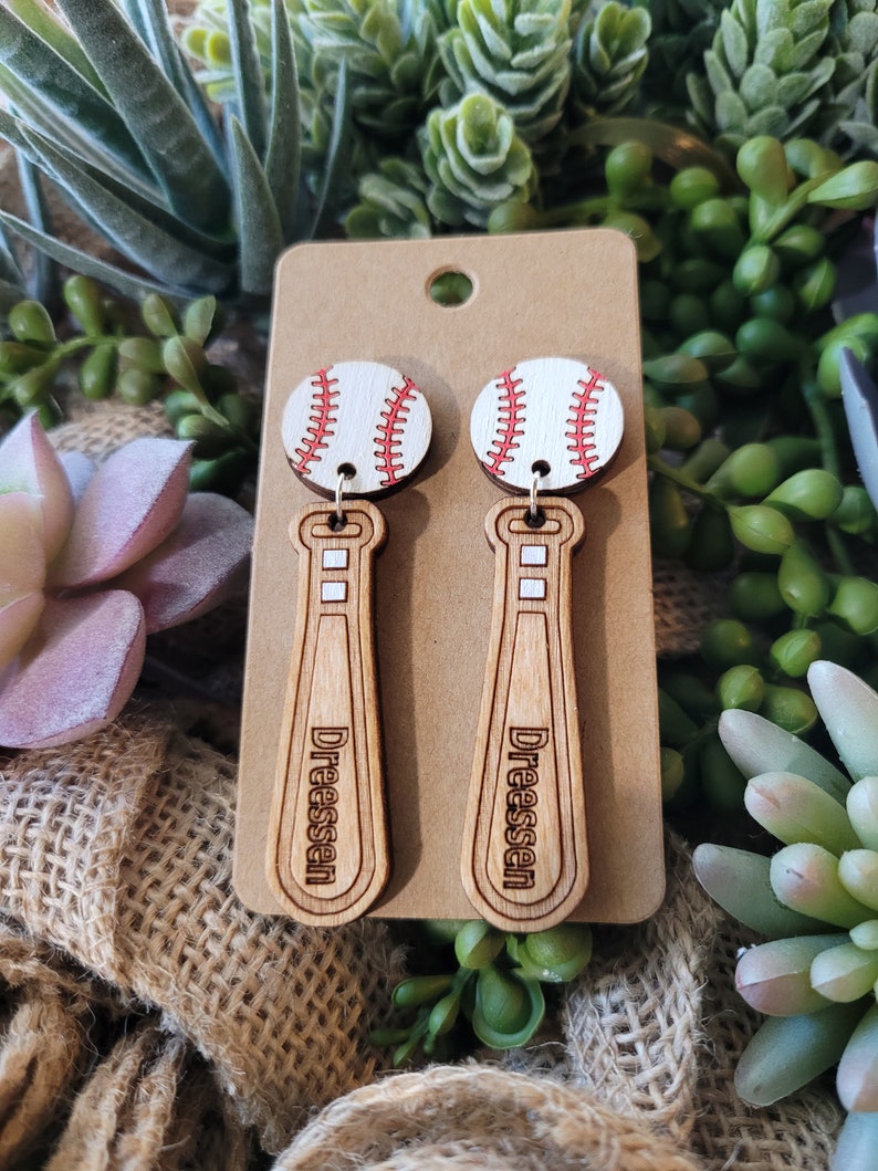 Puede incluir: Un par de pendientes colgantes con una pelota de b&eacute;isbol blanca con costuras rojas unida a un bate de b&eacute;isbol de madera marr&oacute;n claro. El nombre Dreessen est&aacute; grabado en negro en el bate. Los pendientes se presentan en una tarjeta beige.