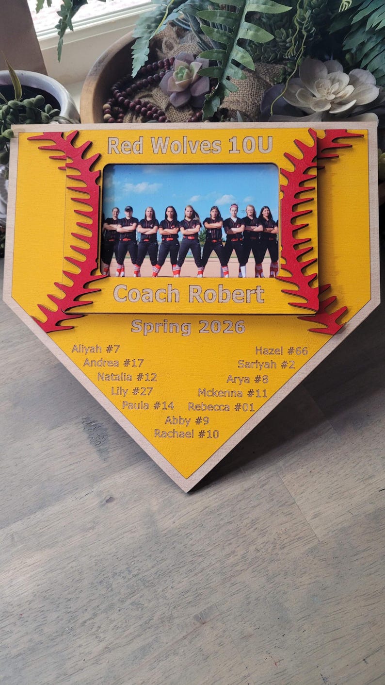 May include: A baseball-shaped wooden plaque with a photo of a softball team, framed in yellow with red stitching. The text "Red Wolves 10U", "Coach Robert", and "Spring 2026" are visible, along with player names and numbers.