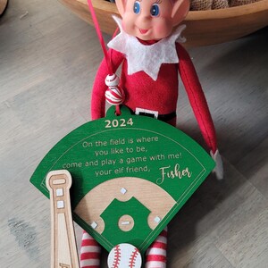May include: A green and brown wooden baseball field ornament with a red and white striped elf leg and a baseball. The ornament has the text "2024", "On the field is where you like to be, come and play a game with me! Your elf friend, Fisher" and "Amari".