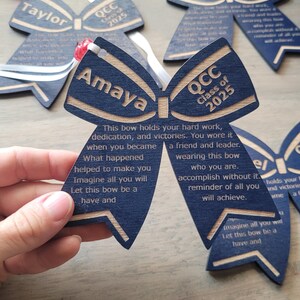May include: Blue and gold wooden bow with the text "QCC Class of 2025" and the name "Amaya". The bow has a message about hard work, dedication, and victories.