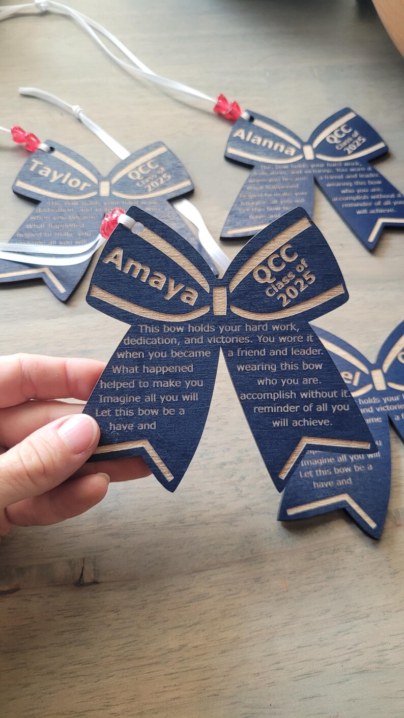 May include: Blue and gold wooden bow with the text "QCC Class of 2025" and the name "Amaya". The bow has a message about hard work, dedication, and victories.
